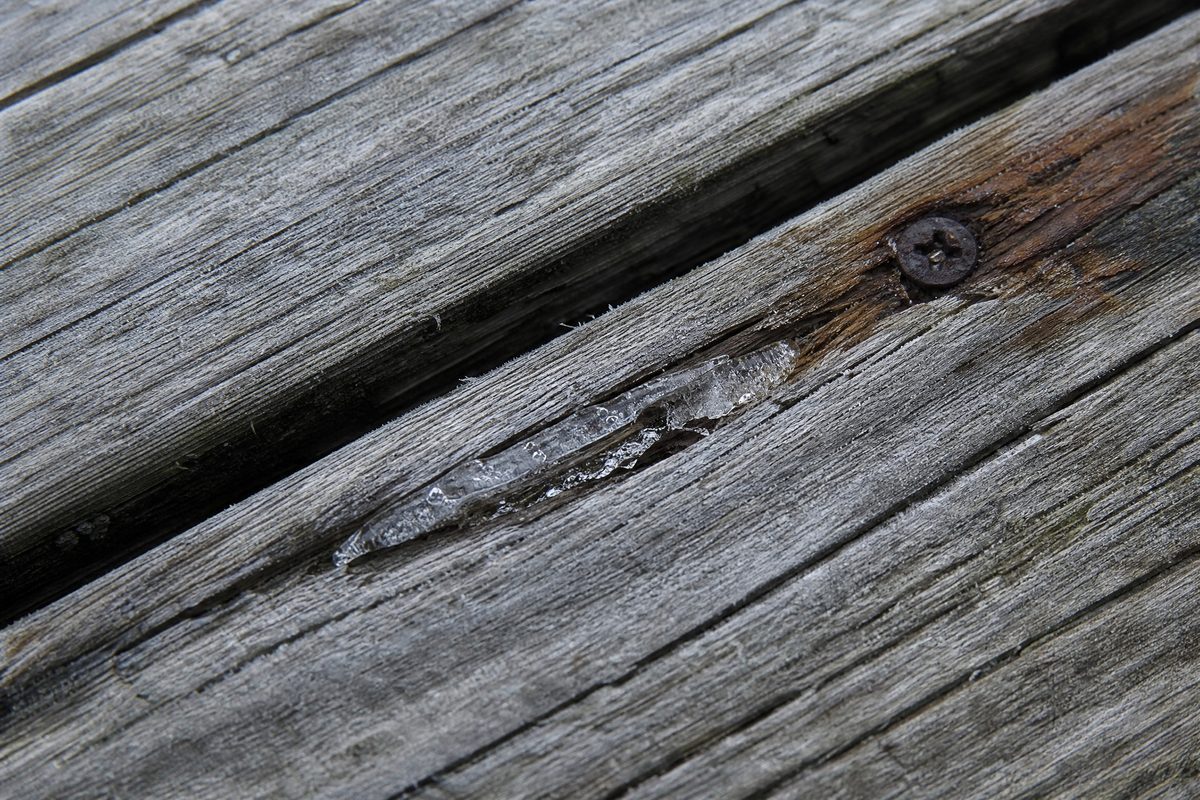 Close-up of weathered grey deck boards with deep splits, raised grain and ice wedged inside a crack
