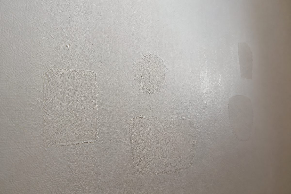 Interior wall in raking light showing repaired patches flashing through the finish coat as duller areas with visible outlines