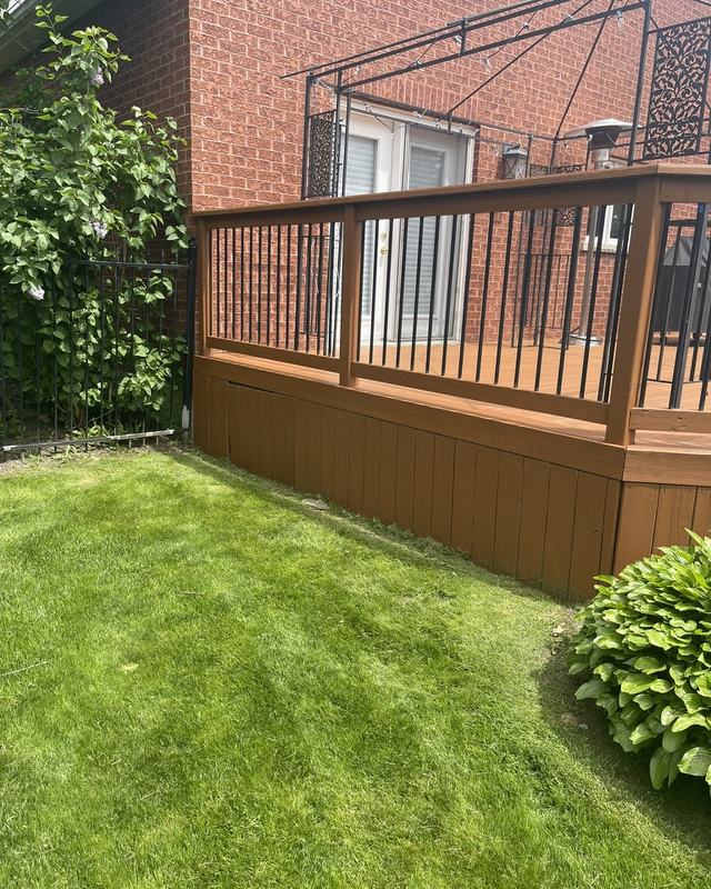 Stained raised deck with black railing attached to a red brick two-storey home, hosta bed and mown lawn below