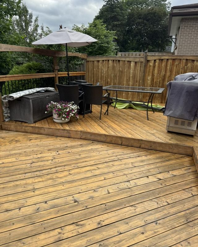 Stained backyard deck with patio umbrella, outdoor sofa and dining set, board fence and mature trees behind