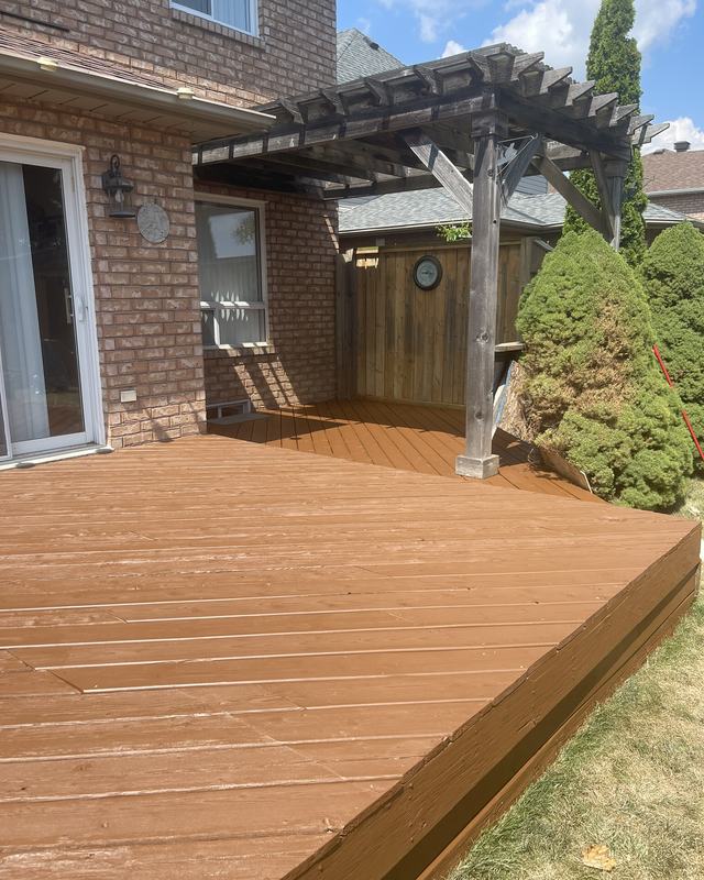 Ground-level deck stained in a warm cedar tone beneath a timber pergola, brick home and privacy fence behind