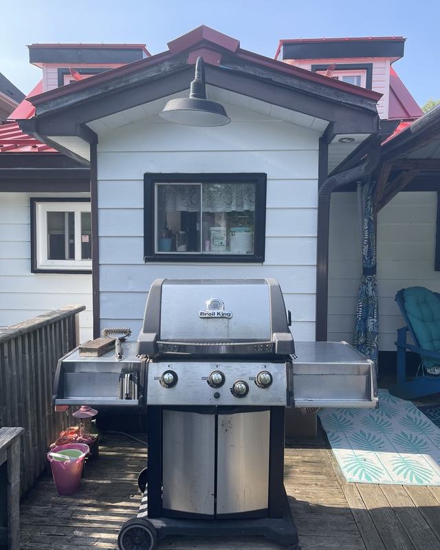 Painted white cottage exterior with a red metal roof, gas barbecue and stained deck in the foreground