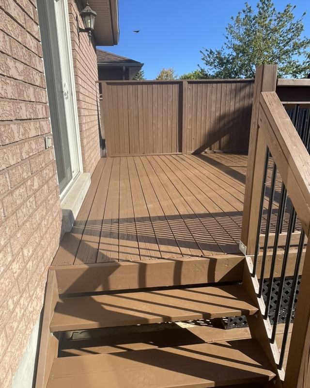 Stained deck with descending stairs, black spindle railing and a cedar privacy panel against a brick wall
