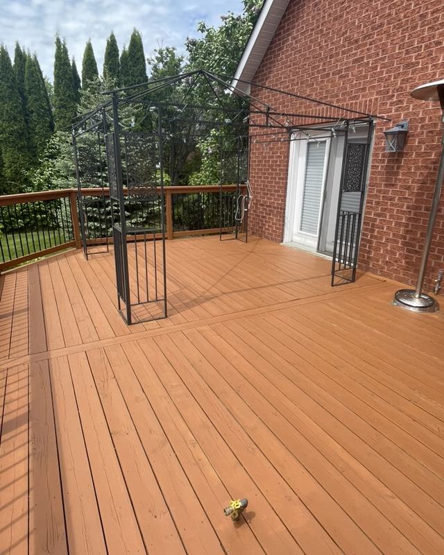 Rear deck of a brick home stained in a warm cedar tone with black railing and a cedar hedge behind
